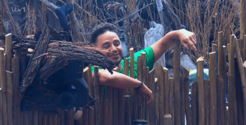 A man in a green shirt smiles behind a wooden fence with a hidden camera.