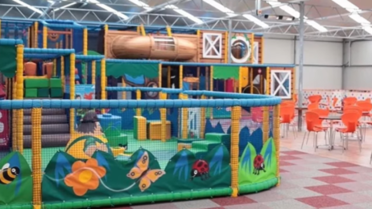 An indoor play area with climbing structures, slides, and nets, next to tables and orange chairs.