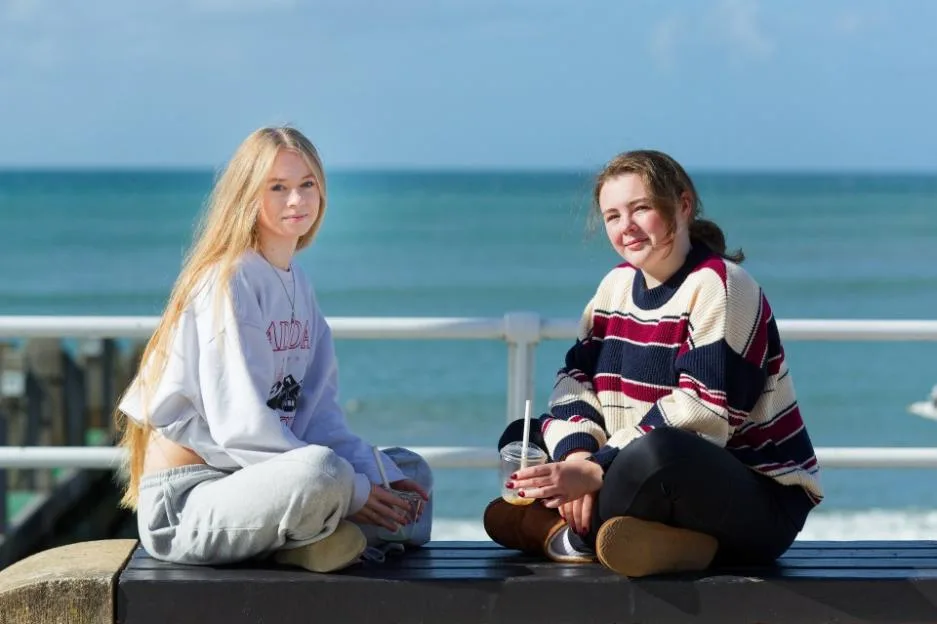 Two university students sitting on a bench by the Aberystwyth seafront, holding soft drinks.