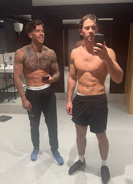 Two shirtless men, one with chest tattoos, stand in a room, with one taking a mirror selfie.