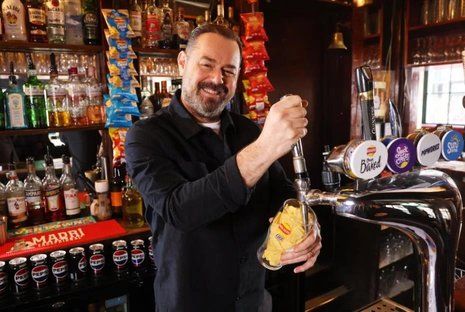 Danny Dyer pulls a pint glass filled with crisps from a beer pump behind a pub bar.