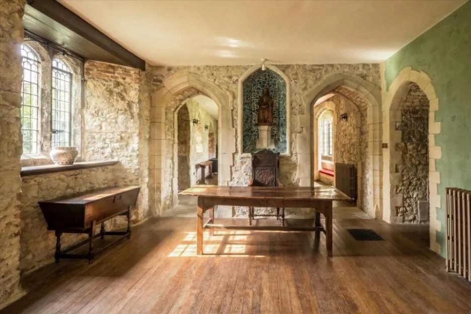 Interior view of a room with exposed stone walls, wooden floors, and arched doorways, featuring a wooden table and chair in the center.
