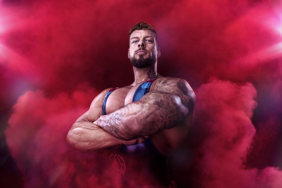 Nick Eagle, a muscular man with tattoos, wearing a blue and red outfit, with arms crossed against a red, smoky background.