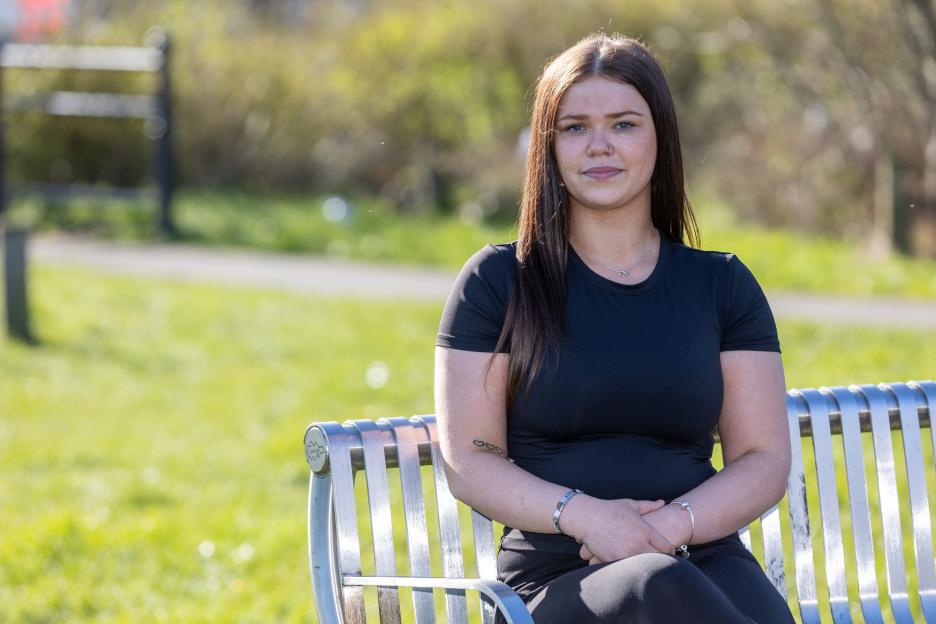 Bobbie Goodman, a young woman with long dark hair, wearing a black t-shirt, sits on a metal slatted bench outdoors.