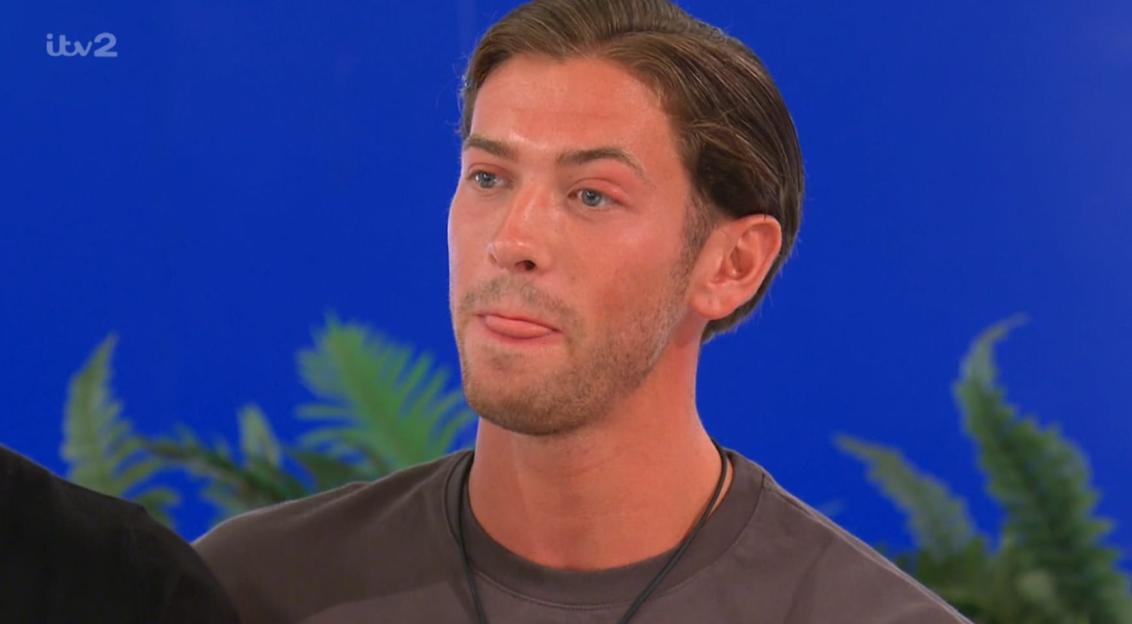A young man with light brown hair, blue eyes, and light stubble on his face, wearing a gray t-shirt and a dark necklace, with his tongue sticking out a little, against a blue background with faint green leaves.