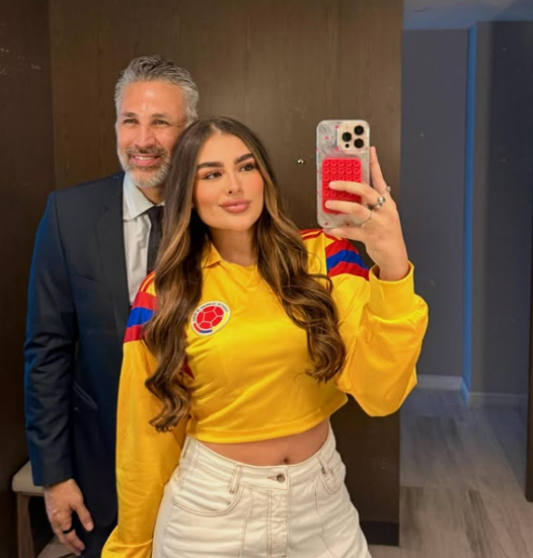 A young woman taking a mirror selfie with an older man smiling in the reflection behind her.