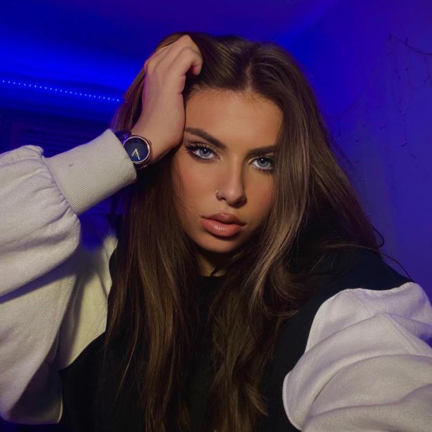 Izzy Sapherson-Moralee, a 22-year-old woman, looking at the camera with her hand in her hair, against a blue background.