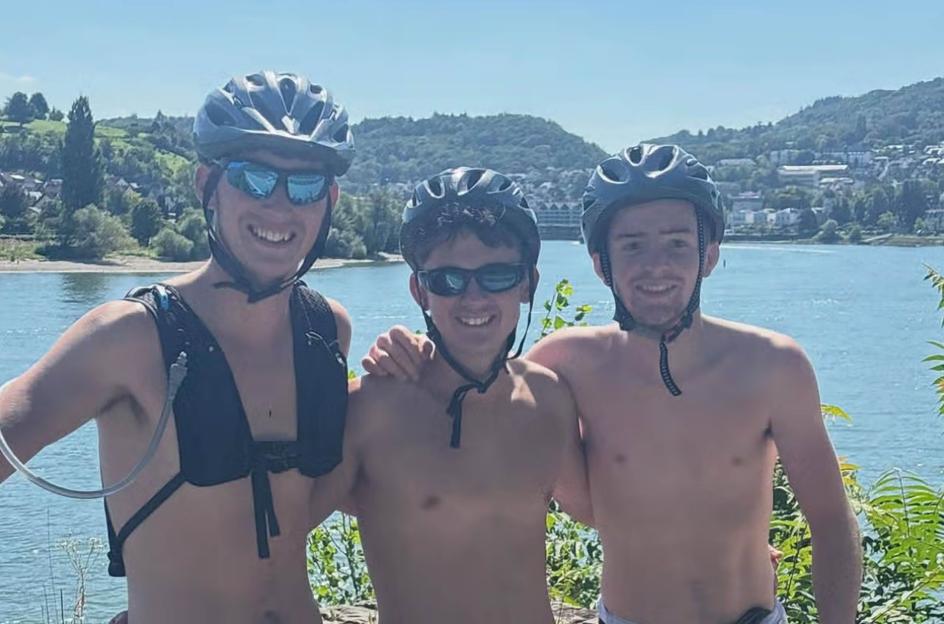 Three shirtless men wearing bike helmets and sunglasses stand by a river.