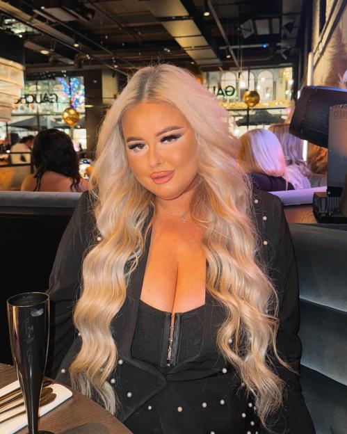 Charly Anne wearing a black blazer and corset, with long blonde hair, sitting at a table with a black flute glass.