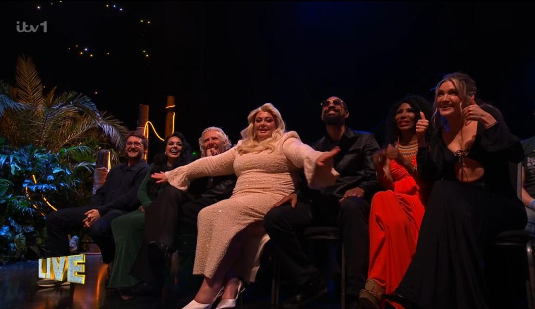 Seven people sit on chairs on a stage in front of tropical foliage, with a "LIVE" sign visible.