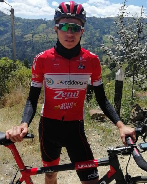 A cyclist in a red and black uniform standing next to his bike with a mountain landscape in the background.