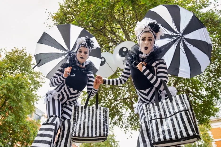 Two stilt walkers in black-and-white striped costumes and makeup hold matching umbrellas and bags in a park.