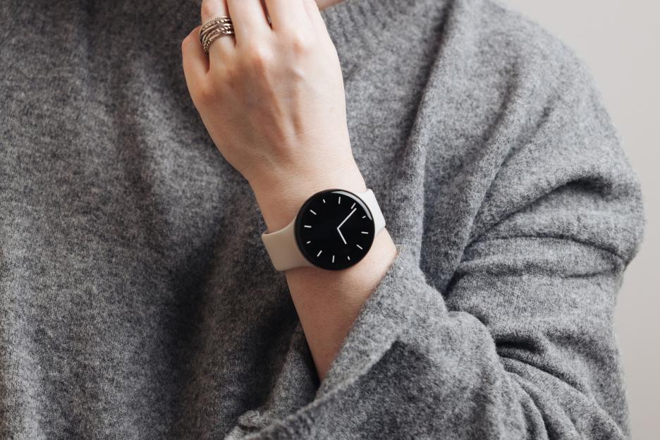 Woman wearing a modern smartwatch with a black circular face and light band on her wrist, with a gray sweater and rings on her finger.