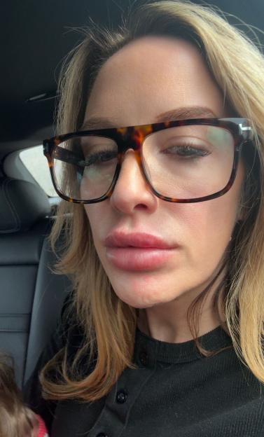 A woman with blonde hair, wearing glasses and a black shirt, looks slightly down.