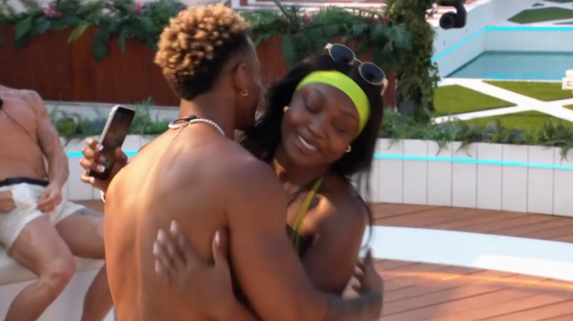 A man with light brown hair and a woman with long dark hair embrace as she smiles.