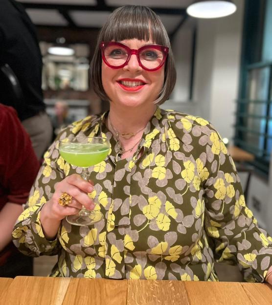 A woman in red glasses and a patterned blouse holding a green cocktail.