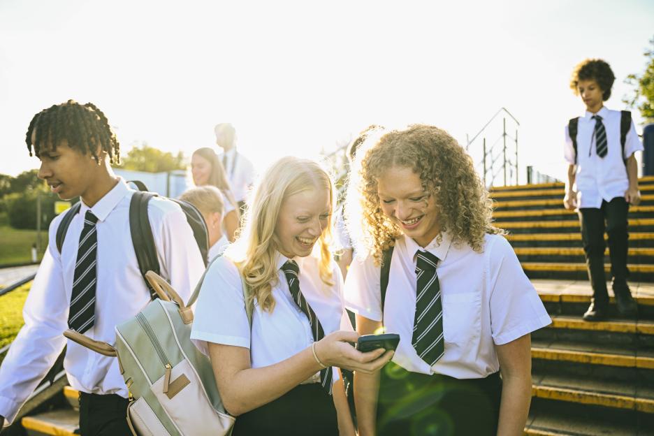 Female high school friends looking at phone before school, students walking past