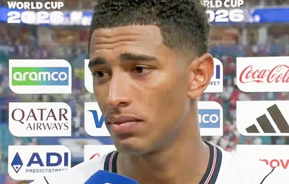 A man with short dark hair and a light complexion in a white shirt with red and black stripes, speaking in front of a blurred background of World Cup 2026 sponsor logos including Aramco, Qatar Airways, and Coca-Cola.