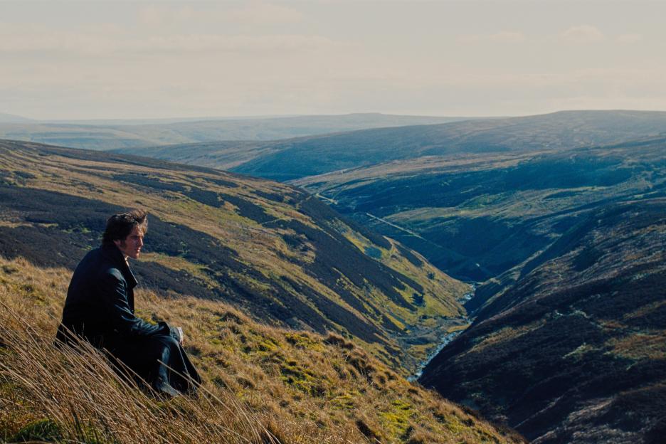 Jacob Elordi in a scene from "Wuthering Heights," sitting on a hill overlooking a valley.