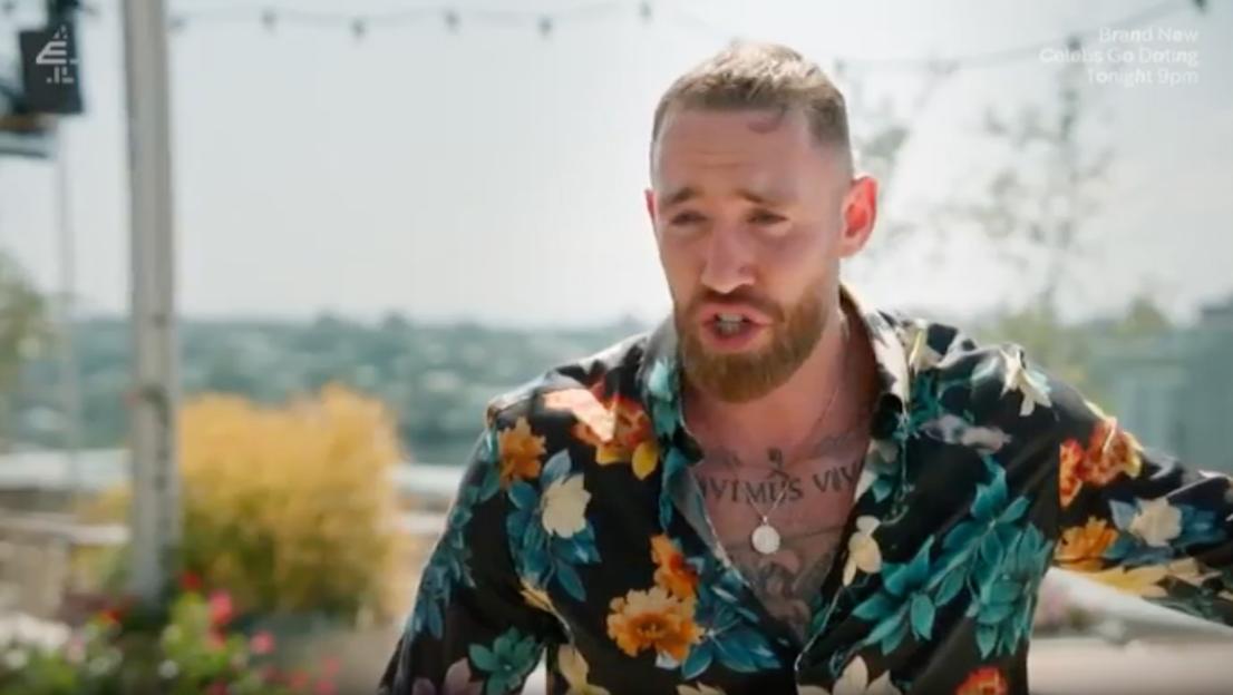 A man in a floral shirt and with a beard, chest tattoos, and necklace is talking.