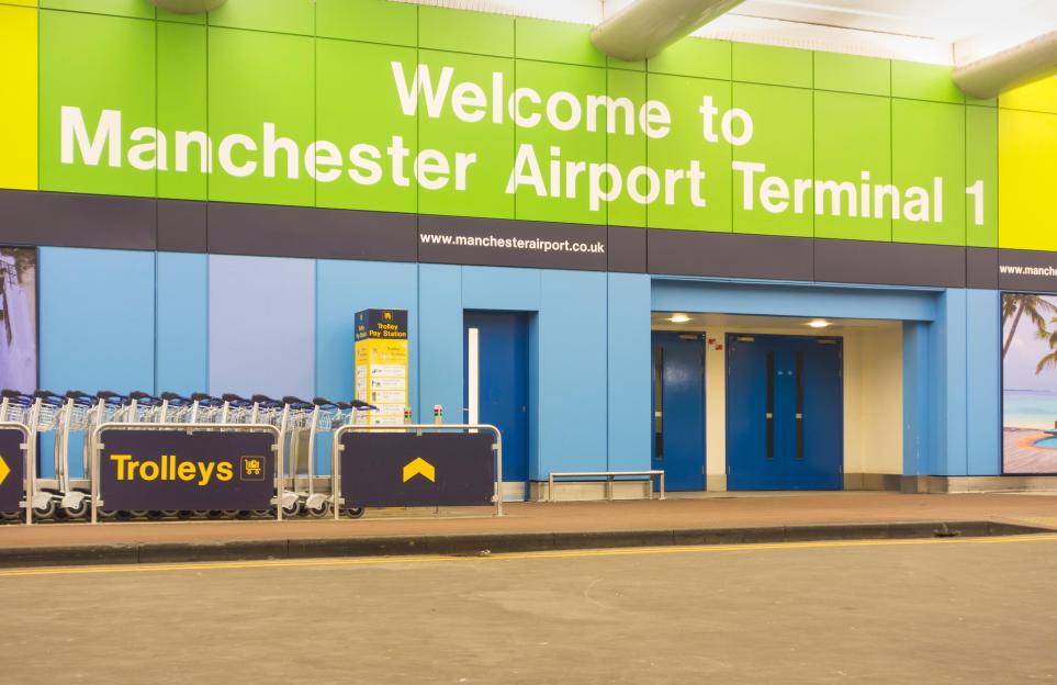 The entrance to Manchester Airport Terminal 1, with a "Welcome to Manchester Airport Terminal 1" sign, blue doors, and a row of trolleys.
