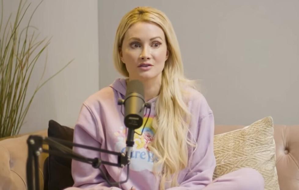 Woman with long blonde hair, wearing a lilac hoodie, speaking into a microphone.