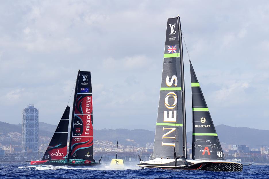 Two America's Cup sailboats, one red and black, the other grey and black, race on the water with a city skyline in the background.