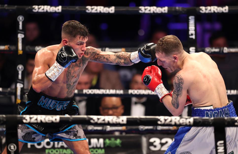 Sam Noakes (left) wearing black shorts and gloves, punches Shaun Cooper (right) wearing blue and white shorts and red gloves, during a boxing match.
