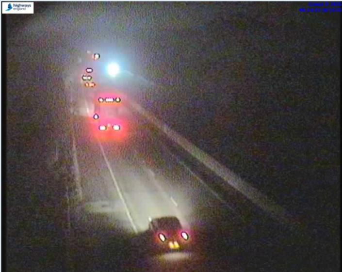 A dark, grainy overhead view of a multi-lane highway at night, with several cars whose headlights and taillights are visible.