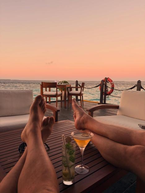 A person's feet and legs resting on a table with cocktails, overlooking the ocean at sunset.