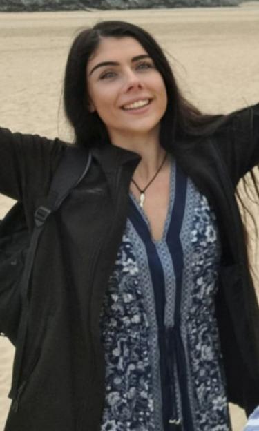 A young woman with long dark hair smiles at the camera, wearing a dark jacket over a blue and white patterned dress.