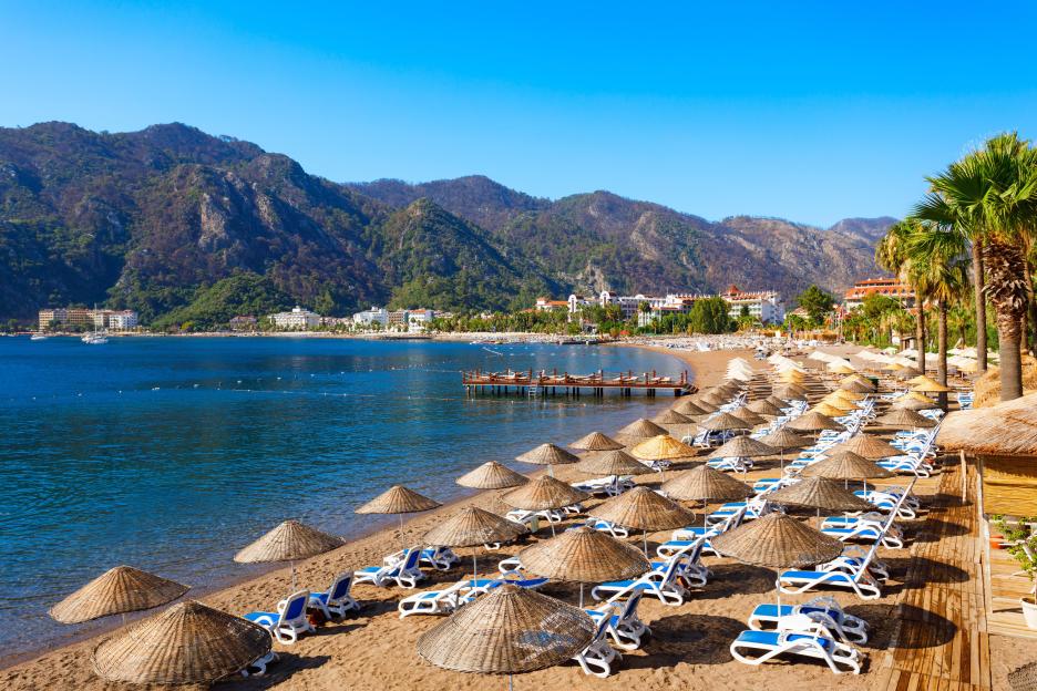 Icmeler main city beach in Turkey with straw umbrellas and beach chairs, with the city and mountains in the background.
