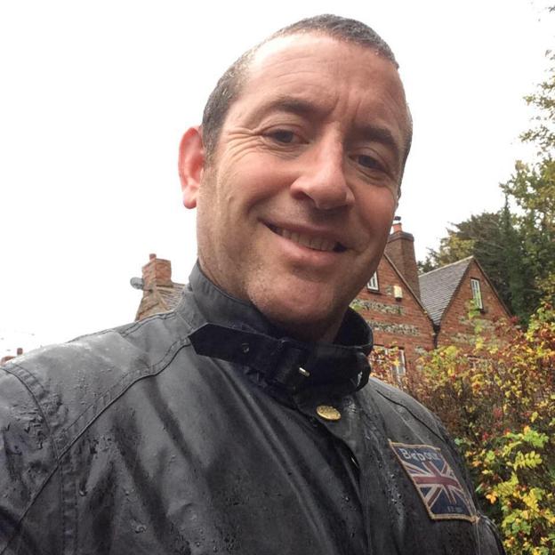 A man smiling and wearing a wet black jacket with a Union Jack patch.