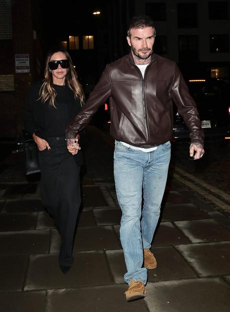 David and Victoria Beckham hold hands while walking together at night, David in a brown leather jacket and jeans, Victoria in black pants and top with large sunglasses.