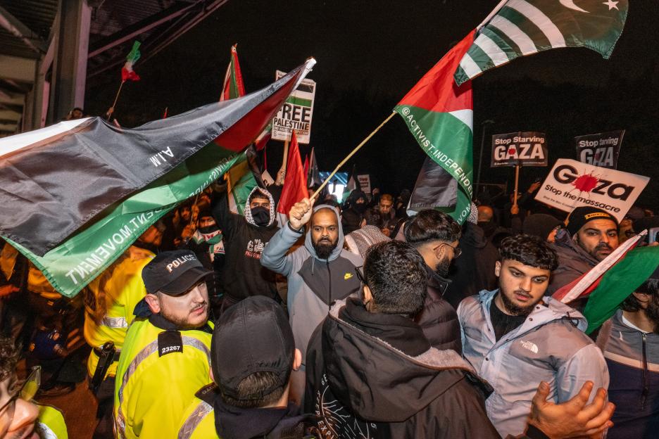 Pro-Palestine activists protest at Villa Park carrying Palestinian flags and signs stating "FREE PALESTINE", "Stop Starving GAZA", and "Stop the massacre" while a police officer observes.