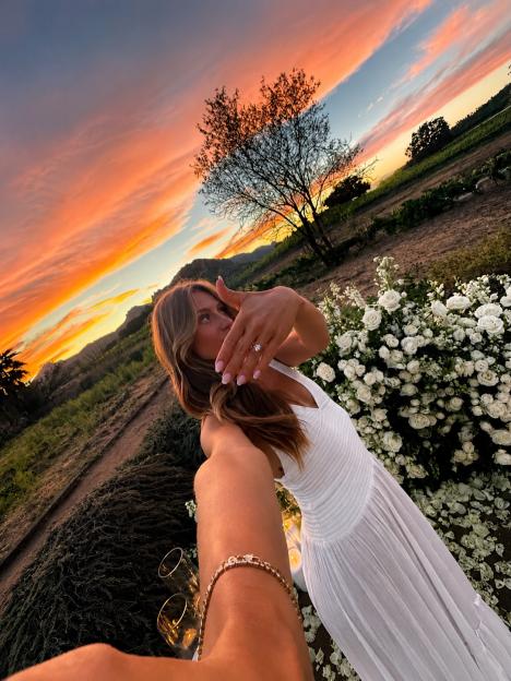 A woman in a white dress shows off her engagement ring at sunset.