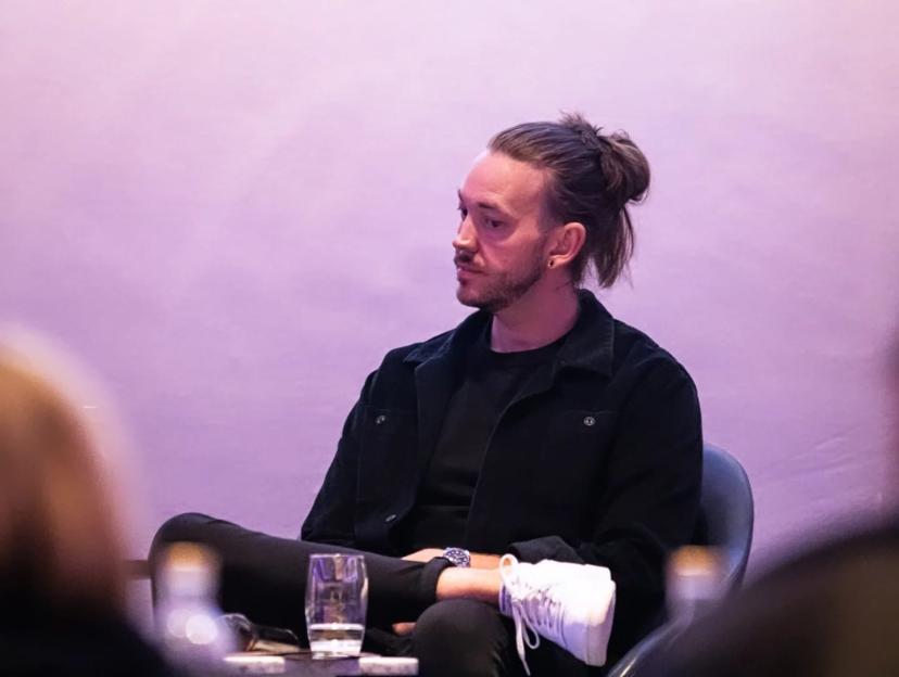 Thomas Delaney seated with his leg crossed, wearing a black shirt and jacket, with his hair in a bun.