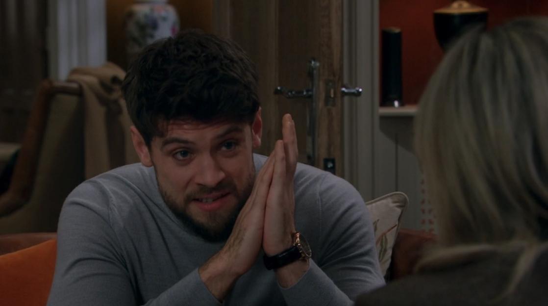 A man with dark hair and a beard, wearing a grey sweater, clasps his hands together in front of him and looks intently at someone out of frame.