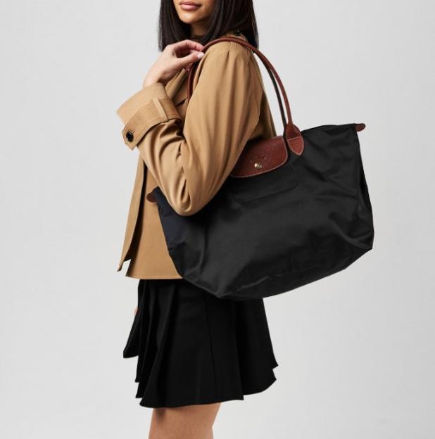 Woman wearing a tan jacket and black skirt carries a black Longchamp Le Pliage Large Tote Bag.