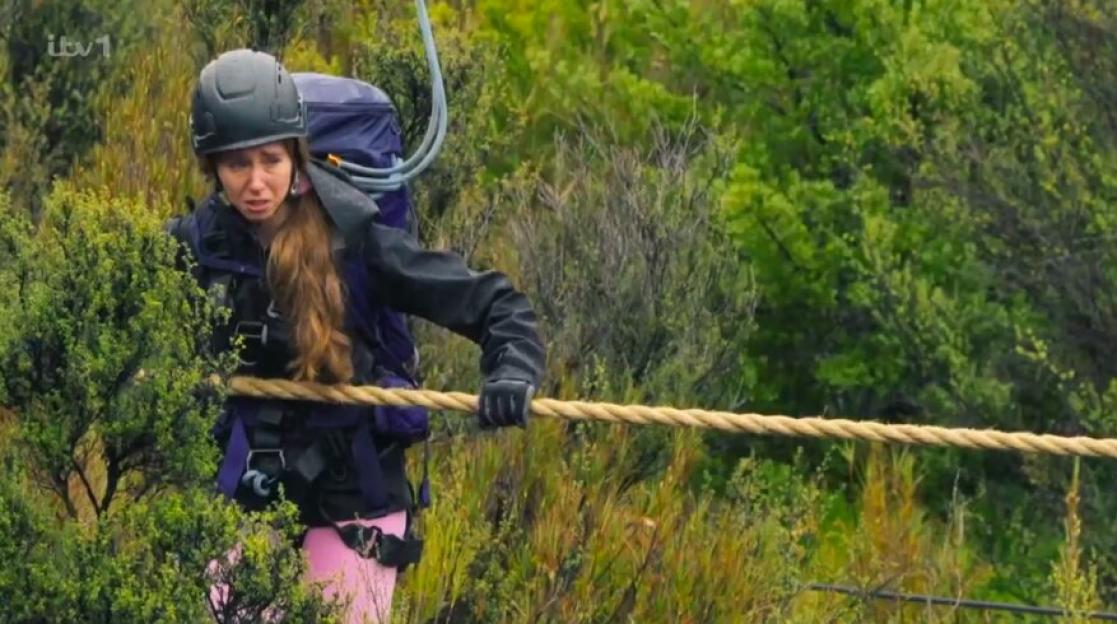 Afton McKeith holding onto a rope, wearing a helmet, backpack, and gloves, looking distressed, surrounded by green foliage.
