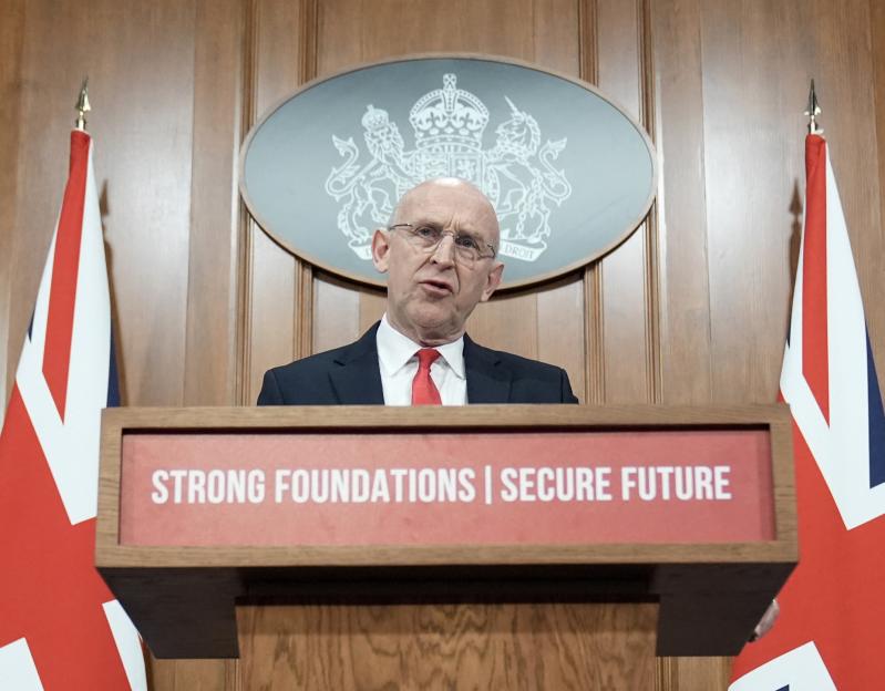 Britain's Defence Secretary John Healey speaking at a podium with two Union Jack flags and a crest behind him.