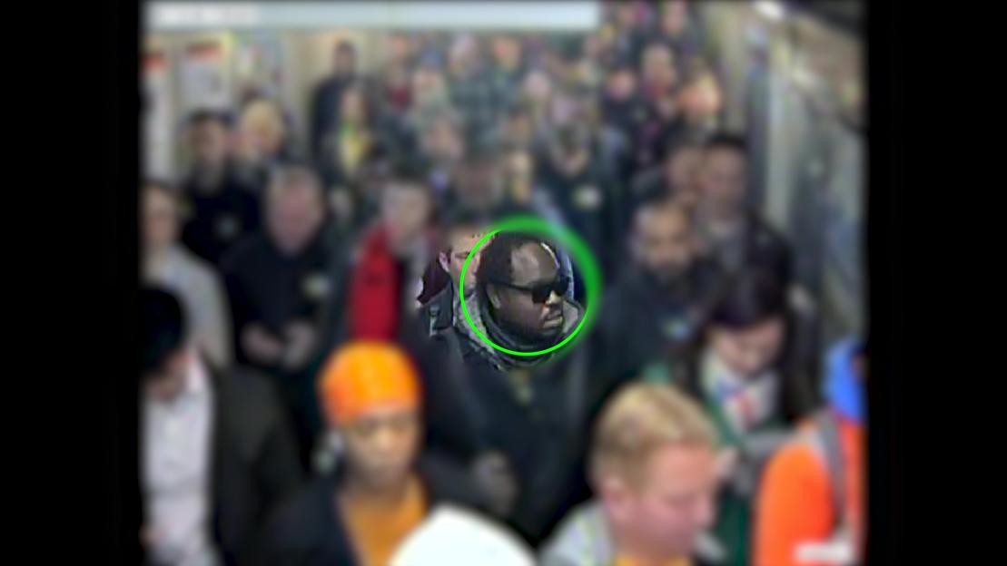 CCTV image of Simon Levy, highlighted in a green circle, wearing sunglasses and surrounded by a blurred crowd at Seven Sisters underground station.