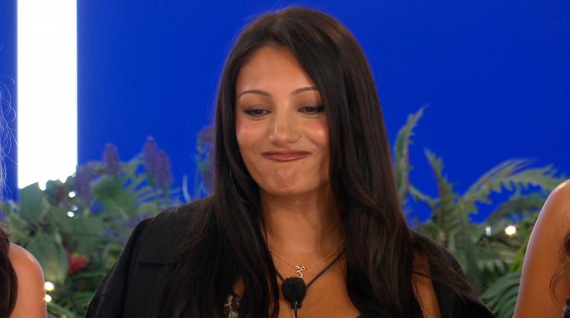 A woman with long dark hair smiles nervously with pursed lips.