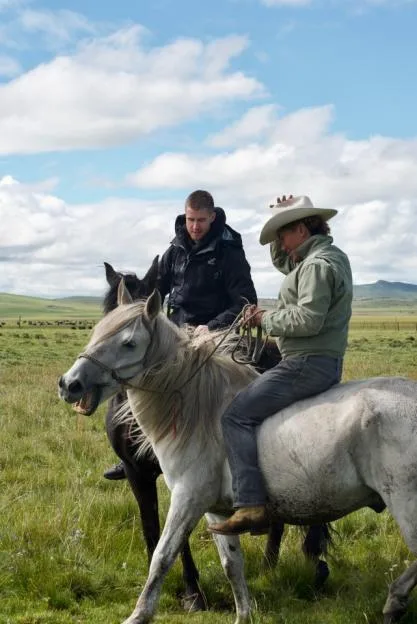 Two men ride horses across a grassy field under a partly cloudy sky.