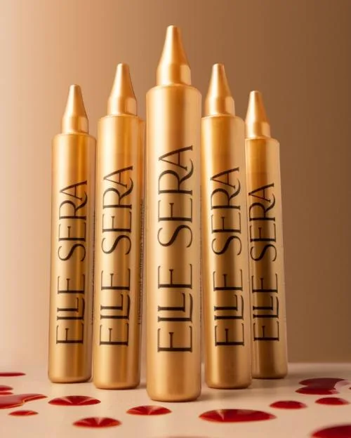 Five gold bottles of ELE SERA serum standing on a light brown surface with red liquid drops scattered around them.
