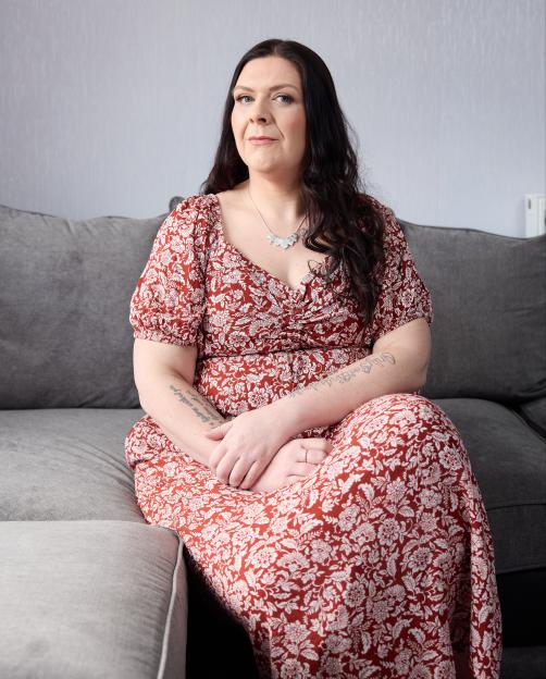 A woman with long dark hair, wearing a red and white floral dress, sitting on a gray sofa.