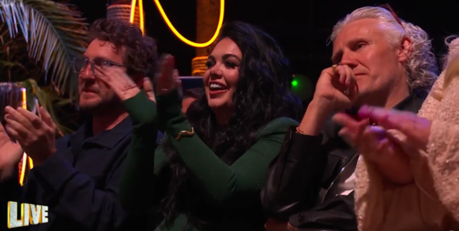 People in an audience clapping, with a woman in the center smiling widely.