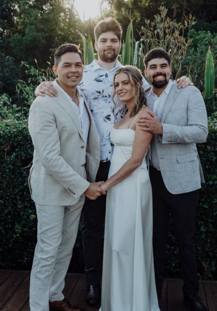 A woman in a white dress holds hands with a man in a beige suit, flanked by two other men.