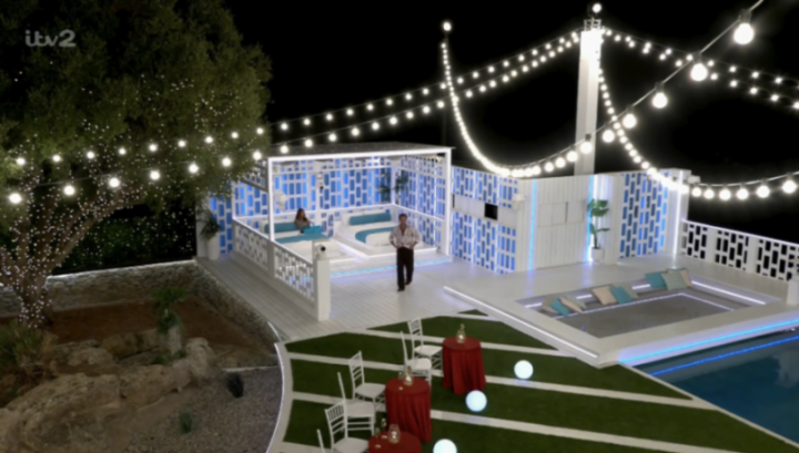 Man walking across a patio with string lights and a pool at night.