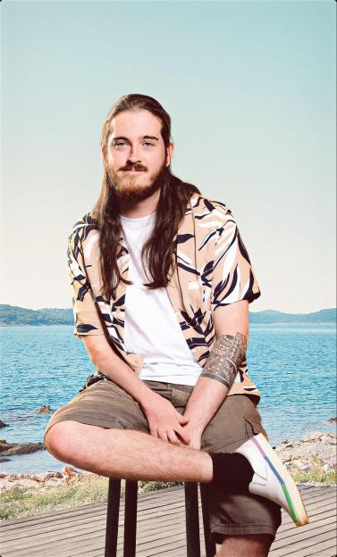 Man with long hair and beard, wearing a patterned short-sleeve shirt, white t-shirt, and shorts, sitting on a stool with a coastal background.